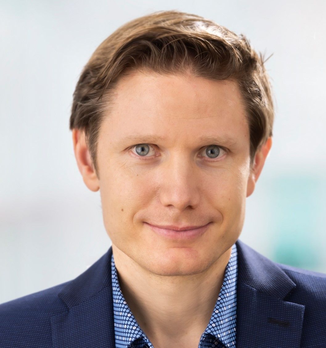 Close-up studio portrait of a person with short, neatly combed hair, facing the camera directly against a neutral, softly blurred background. The person wears a dark blazer over a patterned shirt, with even lighting and sharp focus on the face and upper torso.