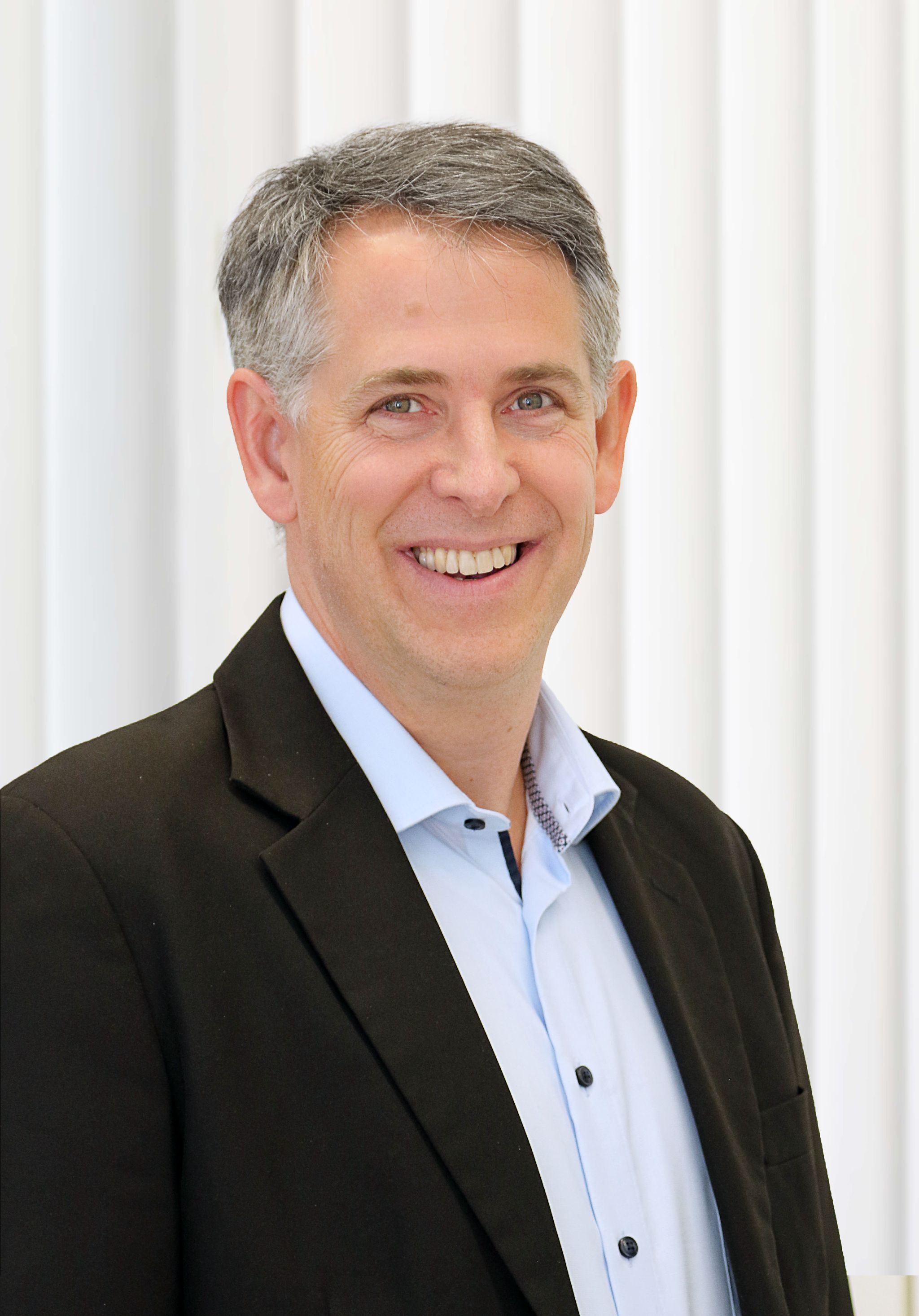 Head-and-shoulders portrait of a person standing in front of a light background with vertical lines, wearing a dark blazer over a light blue button-up shirt. The person has short, graying hair and faces the camera with a slight head tilt. Neutral, evenly lit setting without visible accessories or additional objects.