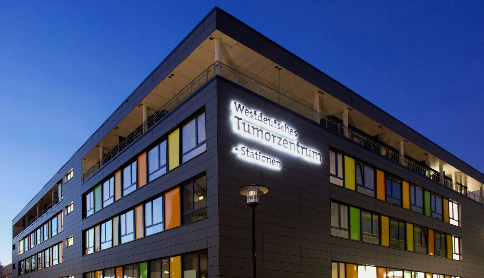 Modern multi-story hospital building at dusk with illuminated signage reading “Westdeutsches Tumorzentrum – Stationen” mounted on the facade. The structure features a clean, angular design with rows of windows, some highlighted by colored panels, and a recessed upper level with a balcony and columns. A streetlamp stands in the foreground against a blue evening sky.