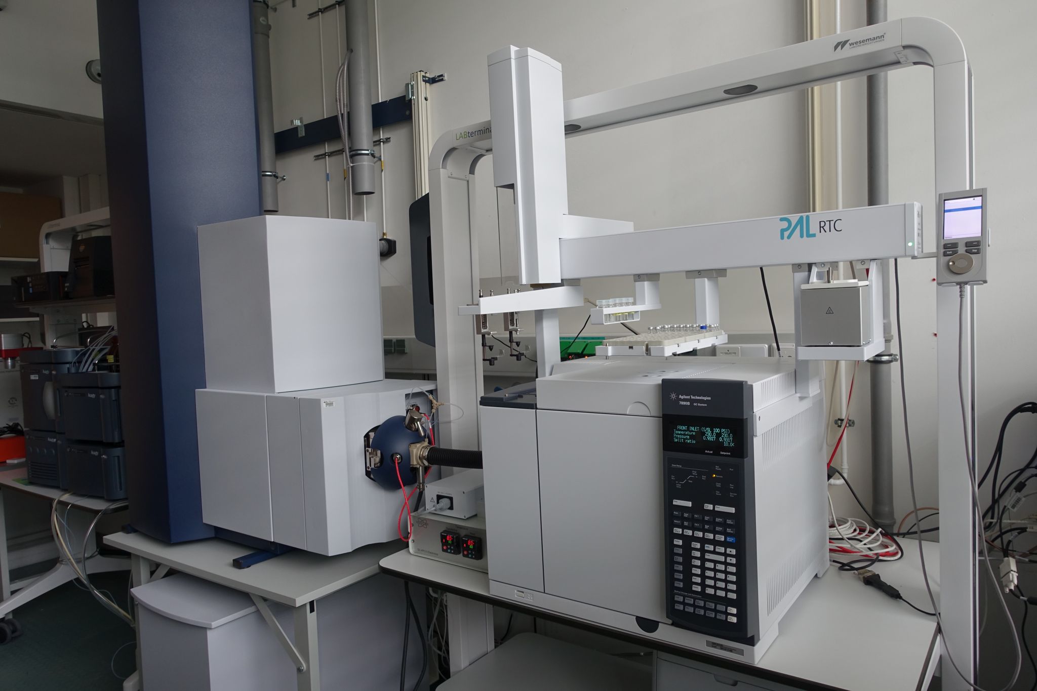 Laboraufbau mit einem Gaschromatographie-System von Agilent (Modell 7890B) und einem automatisierten Probengeber (PAL RTC), verbunden über eine Greif- und Injektionseinheit mit Probenracks. Mehrere Module stehen auf einem Labortisch, mit sichtbaren Leitungen, Steuerdisplay und angeschlossenen Versorgungskomponenten im Hintergrund. Die Anordnung zeigt eine automatisierte Probenvorbereitung und -analyse in einer instrumentellen Analytikumgebung.