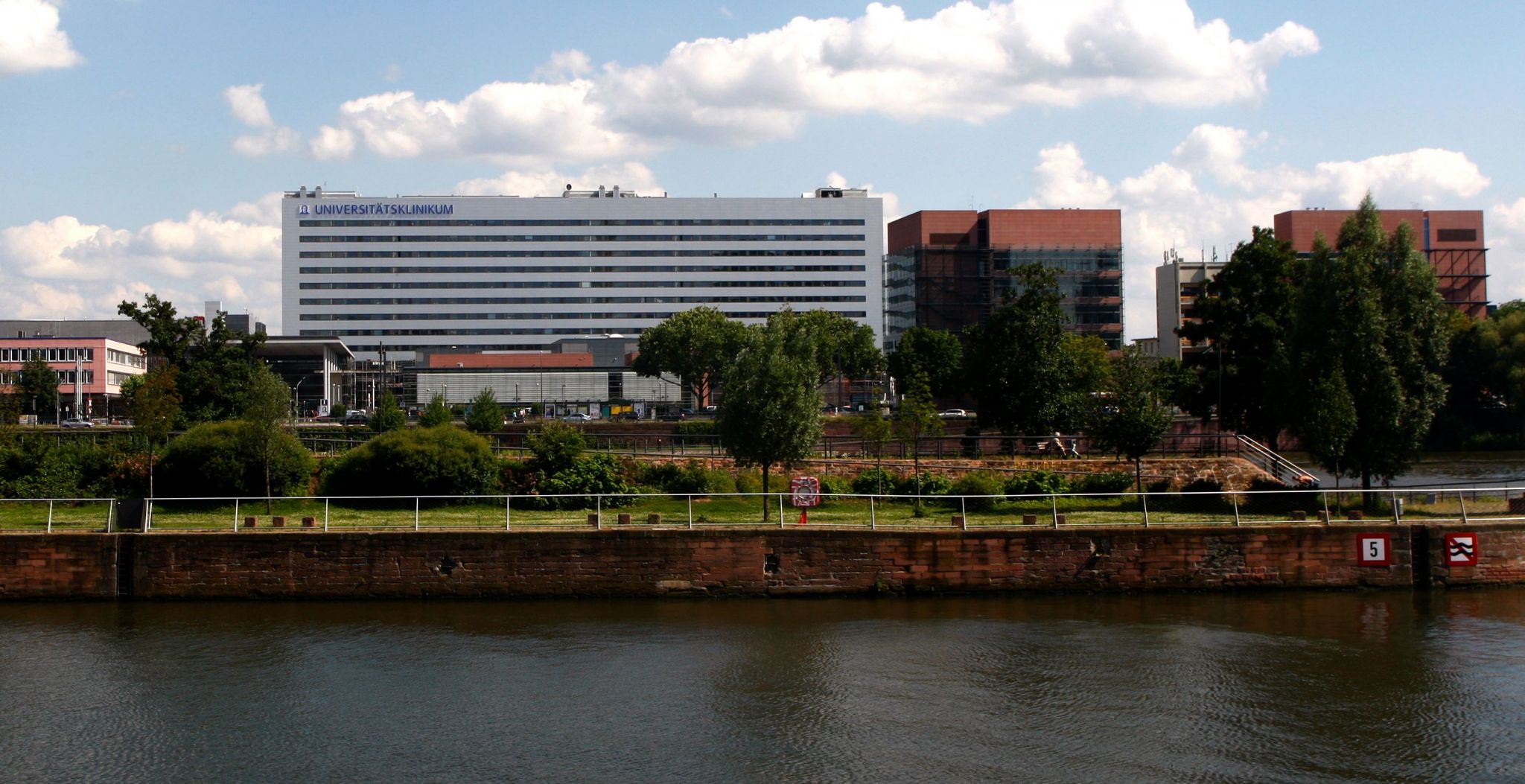 Large university hospital complex beside a नदी or canal, featuring a long multi-story main building with adjacent structures, trees, and a riverside walkway in the foreground. The building façade displays the text “Universitätsklinikum,” with a bright sky and scattered clouds above. The scene presents an extensive medical campus within an urban setting.