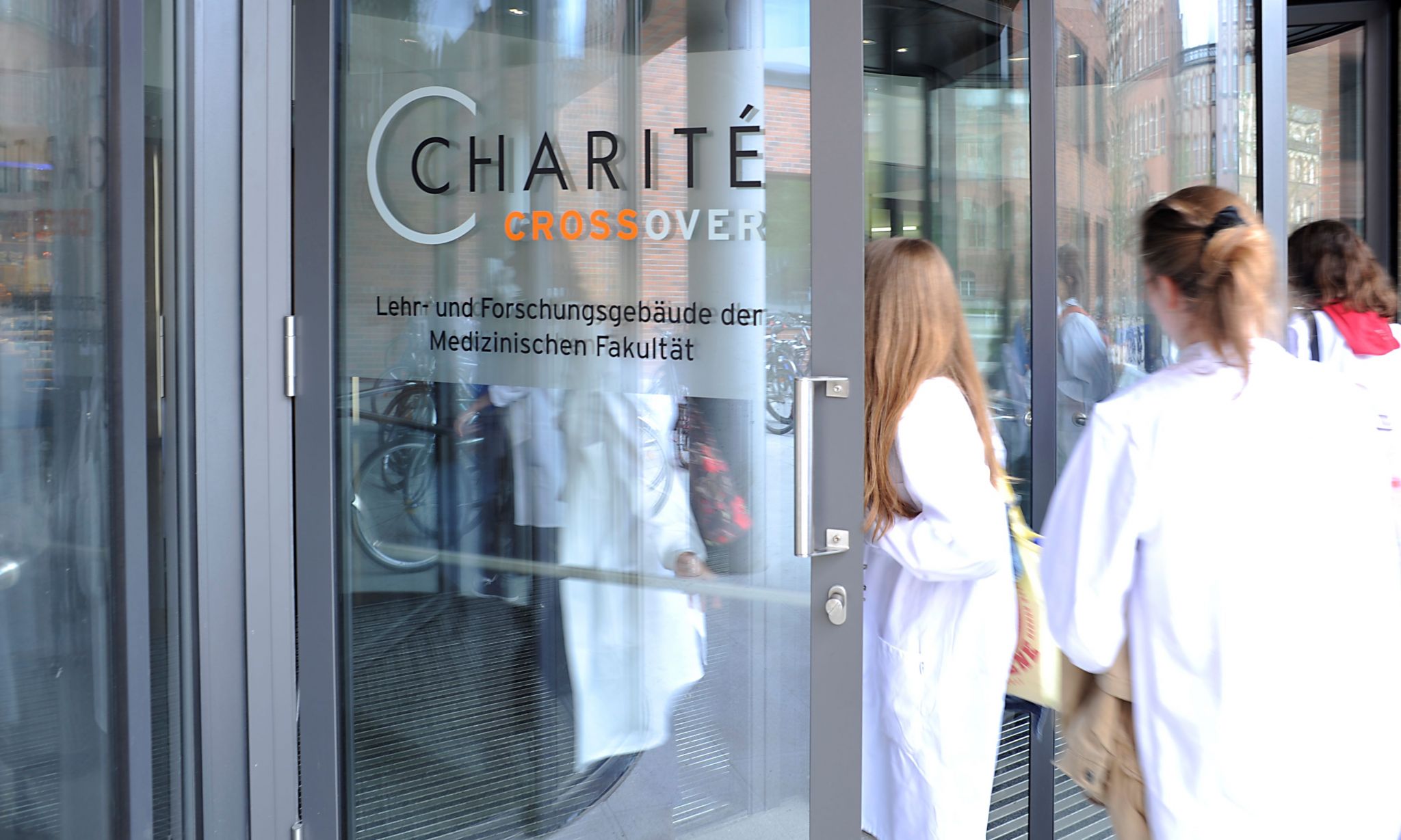 Glass entrance door labeled “Charité Crossover – Teaching and Research Building of the Medical Faculty,” with several people in white coats entering; reflections in the glass reveal bicycles and surrounding buildings.