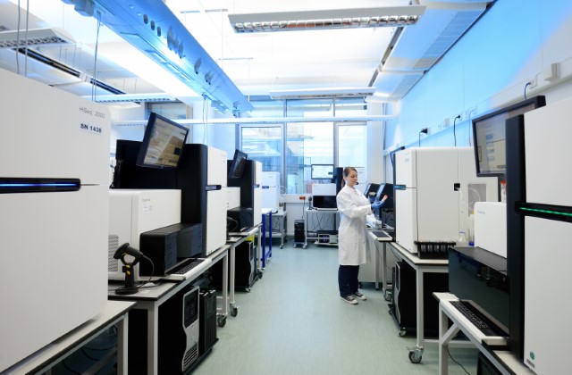 Modern laboratory with several large sequencing instruments arranged on workstations, where a lab-coated person operates one of the systems; monitors and control panels are integrated into the devices, with cables and auxiliary units visible beneath the benches. The setup illustrates a structured high-throughput genomics environment with parallel sample processing in a bright, well-equipped room.