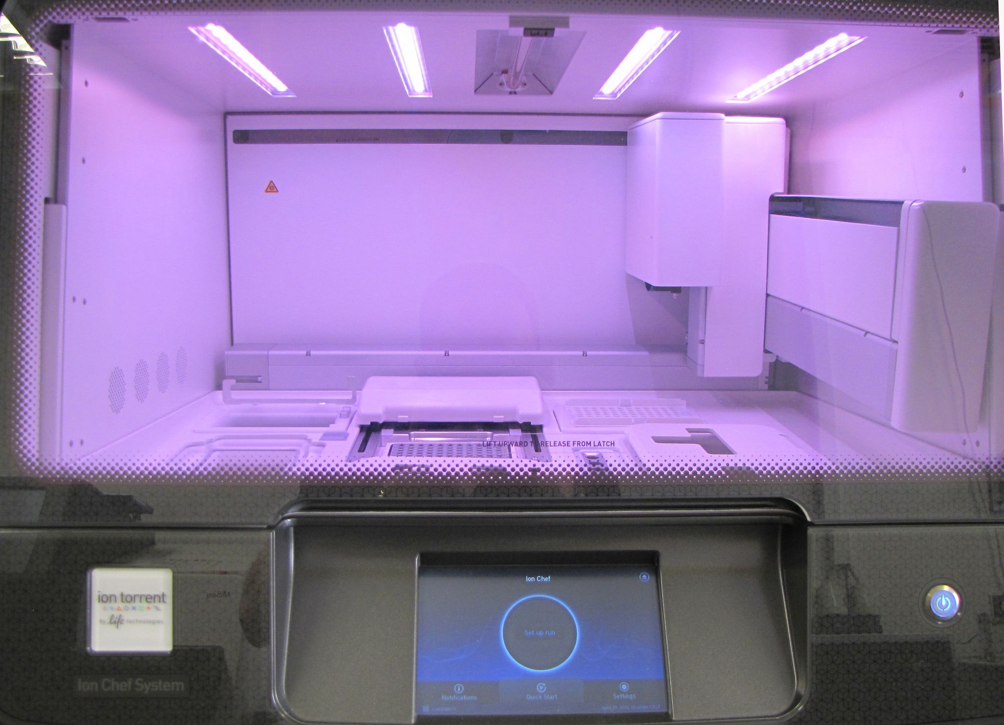 Innenraum eines automatisierten Laborgeräts (Ion Chef System) mit geöffneter Kammer, beleuchtet durch violette LEDs, in der sich eine flache Arbeitsplattform, Probenhalterungen und ein mechanischer Greifarm zur Handhabung von Reagenzien und Kartuschen befinden. Eine transparente Frontscheibe erlaubt den Blick auf die Arbeitsfläche mit mehreren Einsätzen und einem mittig positionierten Modul.