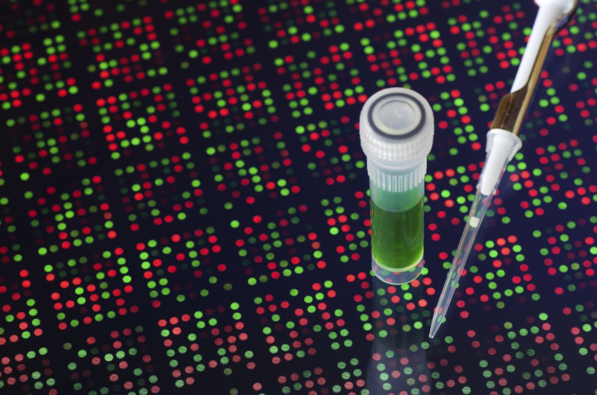Test tube containing green liquid and a pipette tip placed on a surface covered with many colored dots resembling a DNA microarray or genetic data visualization. The composition highlights the link between laboratory samples and the analysis of genetic information.